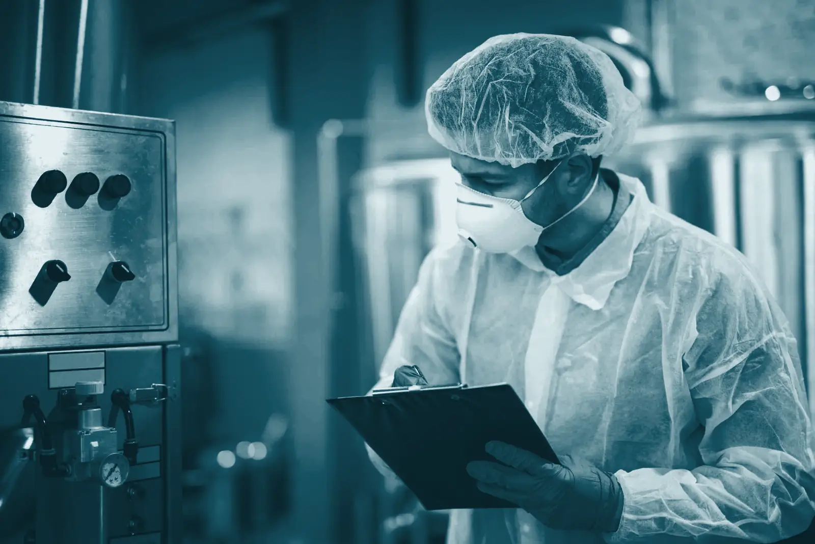 A processing technician in protective gear checks a clipboard beside production equipment